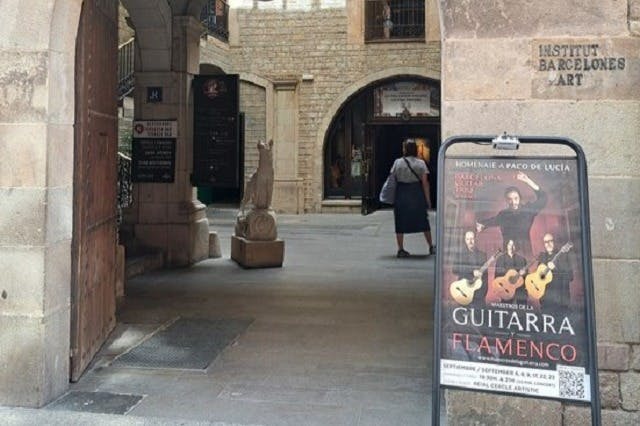 Une personne marchant dans une cour près d'un panneau annonçant un concert de guitare flamenco, avec de vieux bâtiments en pierre en arrière-plan.