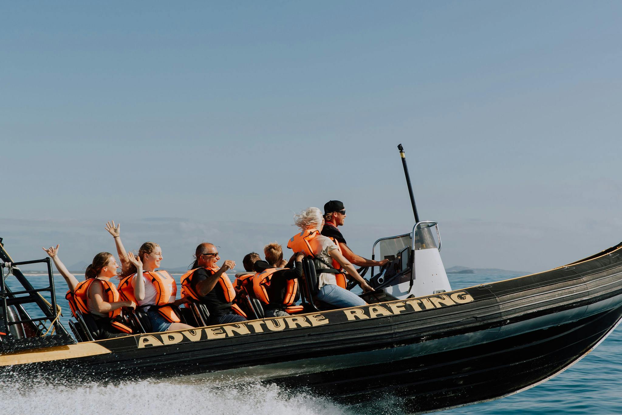 Люди в оранжевых спасательных жилетах ездят на черном быстроходном катере под названием «ПРИКЛЮЧЕНЧЕСКИЙ РАФТИНГ» по чистой голубой воде.