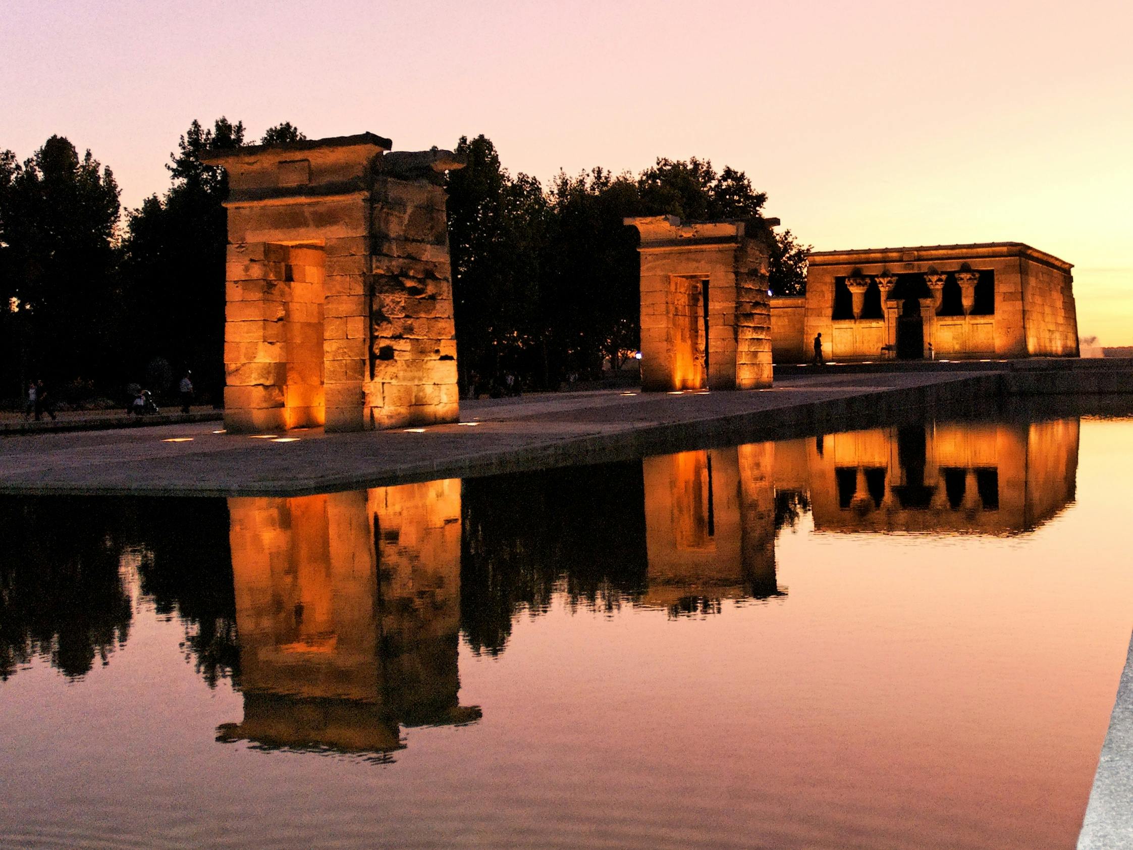 在黄昏时分，被温暖的灯光照亮的古老石头结构反射在平静的水面上，背景是树木。