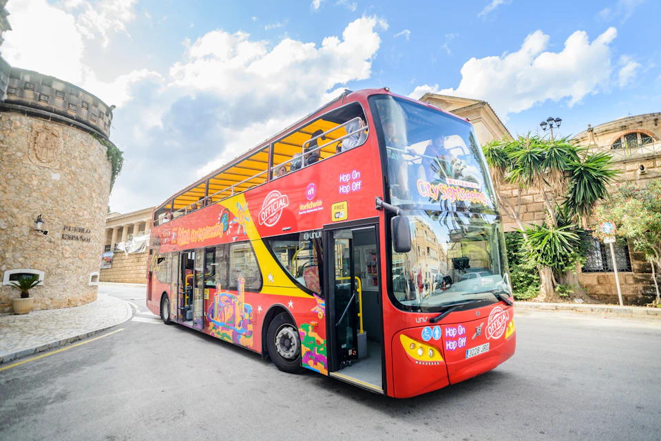 A red double-decker sightseeing bus with an open top is parked on a street in a sunny location.