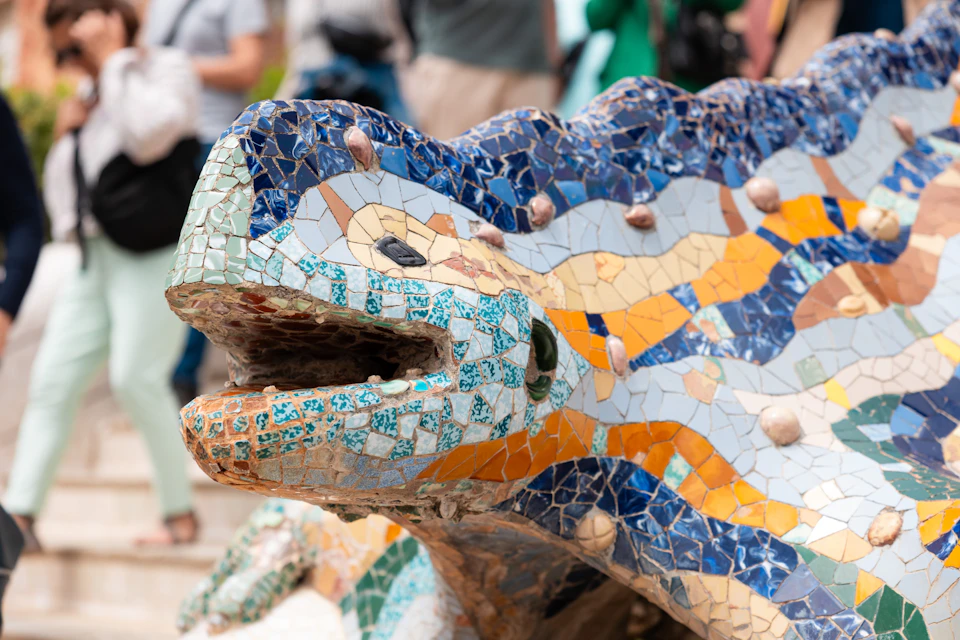 A close-up of a colorful mosaic sculpture of a lizard with a textured surface and a background of blurred people.