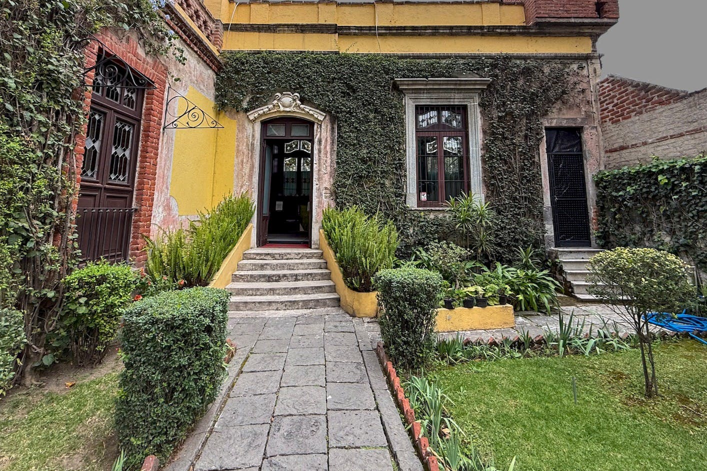 Un pati amb un camí de pedra que condueix a un edifici groc amb parets cobertes d'hiedra, finestres i una petita escala a una entrada.