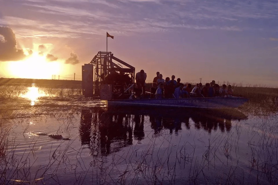 A group of people on an airboat ride at sunset, reflected on a water surface with marshland vegetation.