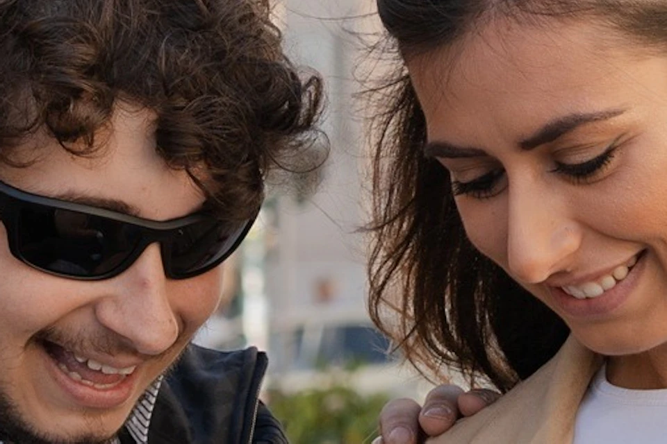 Two individuals outdoors, one pointing at a phone held by the other, with a cityscape and dome in the background.