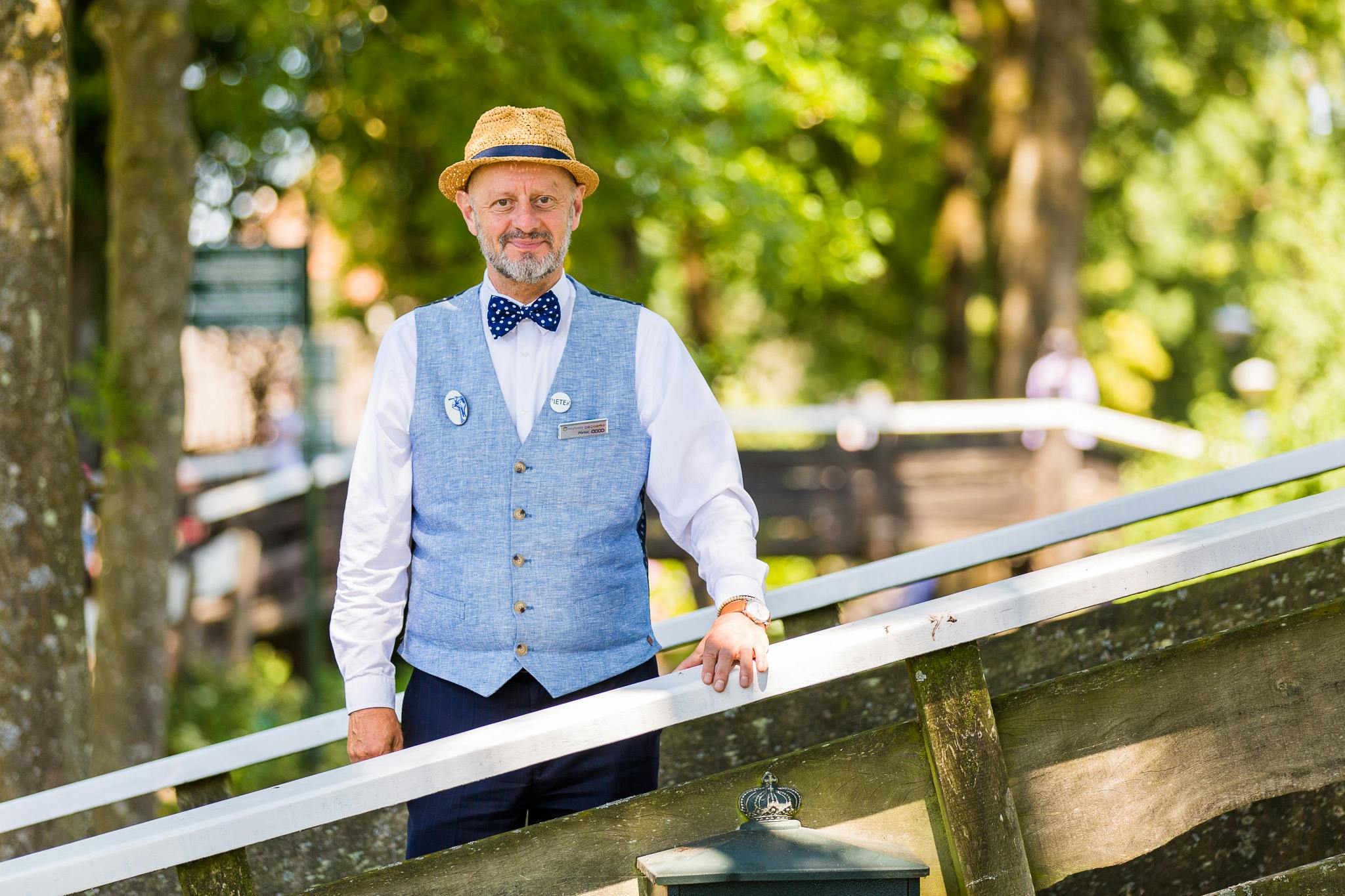 An older man in a straw hat, blue vest, and bow tie stands outdoors, smiling, with trees blurred in the background.