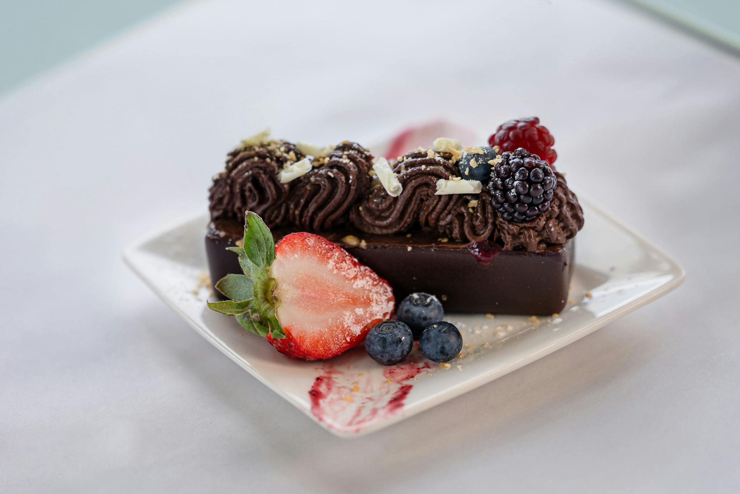 A rectangular chocolate dessert topped with piped chocolate mousse, berries, and white chocolate shavings on a white plate.