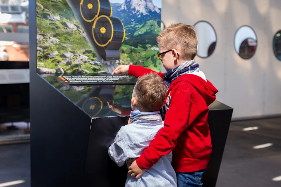 Two boys are looking at and pointing to an exhibit display inside a room with circular windows. One boy is wearing red.