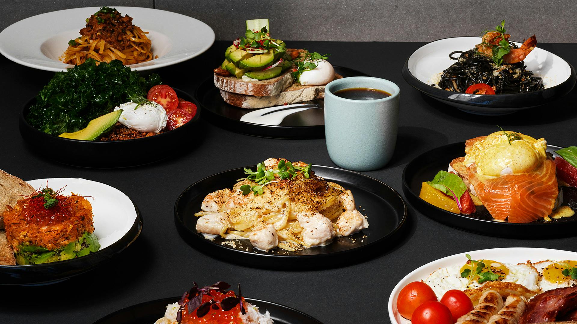 A table set with various dishes including avocado toast, pasta, chicken, and a cup of coffee.