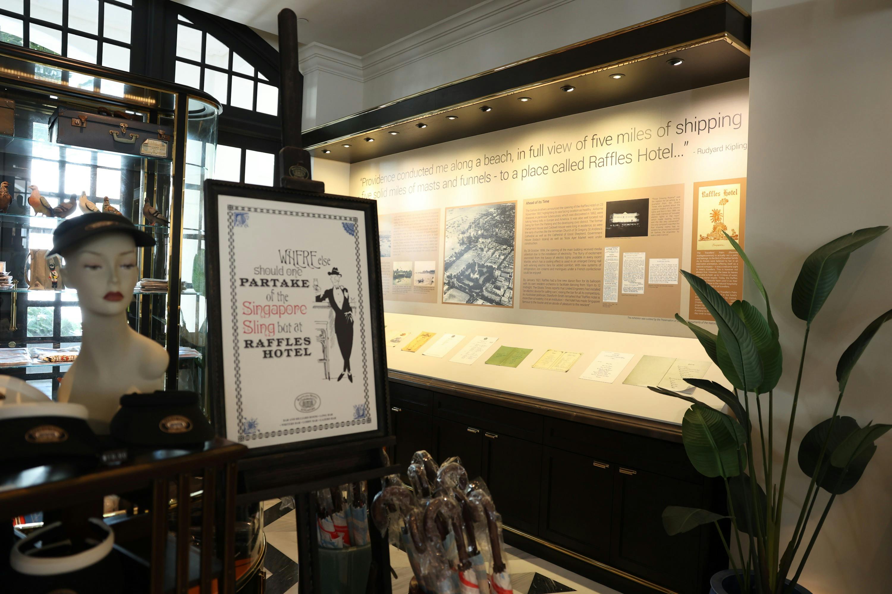 A display in a museum or shop with a mannequin head, a framed poster about Raffles Hotel, and informational panels on the wall.
