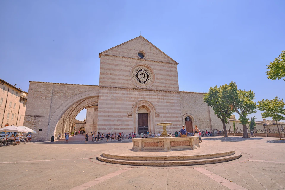 A historic stone church with a rose window and an arched entryway, surrounded by a plaza with a fountain and people.