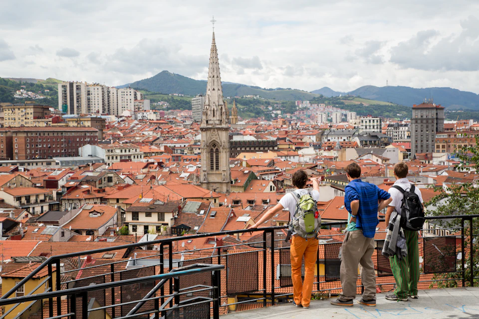 Four people stand on a viewing platform overlooking a city with red-tiled roofs and a tall church spire. Hills are in the background.