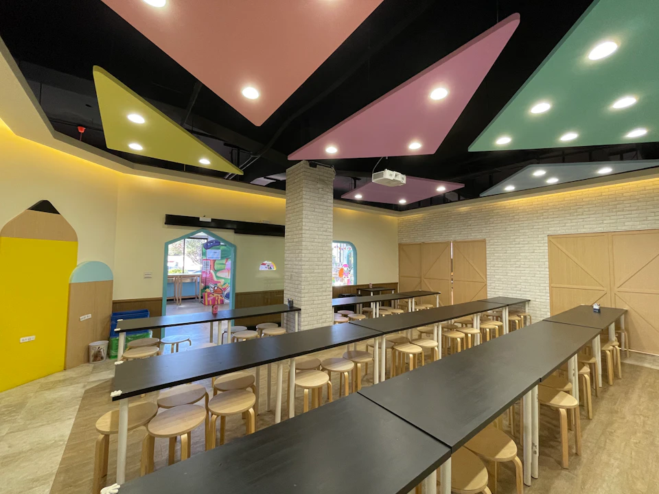 A modern, colorful room with long tables and stools, pastel ceiling panels, and arched doorways leading to another area.