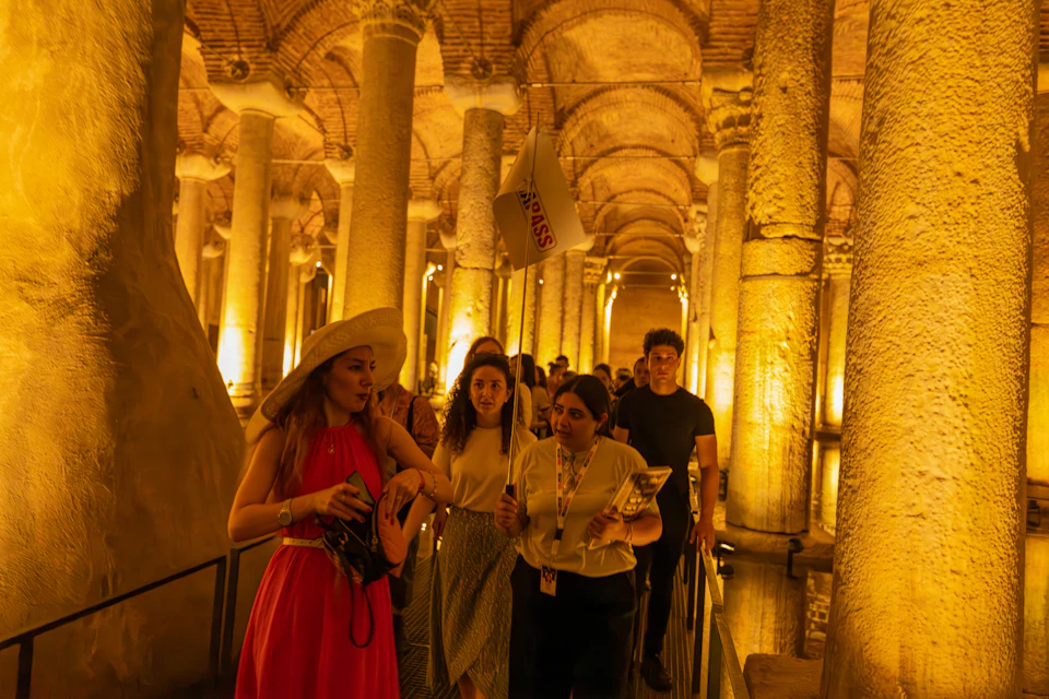 A group of people walking through an ancient, illuminated columned hall. One person holds a sign, possibly leading a tour.