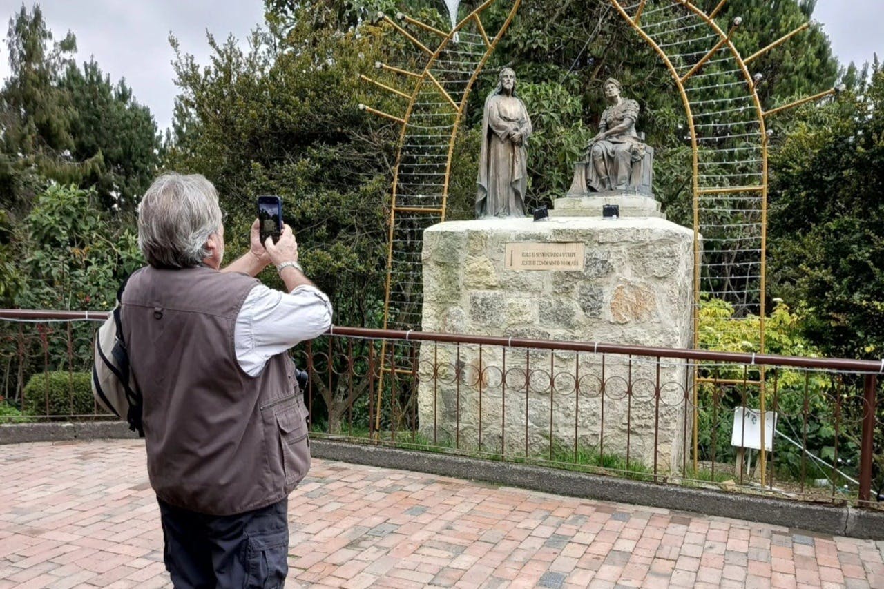 Člověk vyfotí kamenný podstavec se dvěma sochami v parku, obklopený zelení a kovovým obloukem.