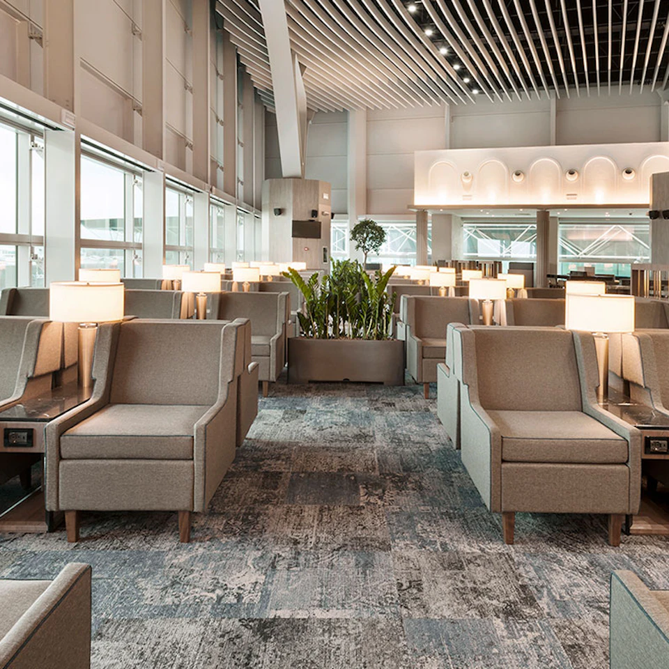 Spacious airport lounge with grey armchairs, small tables with lamps, and large windows. Central plant arrangement.