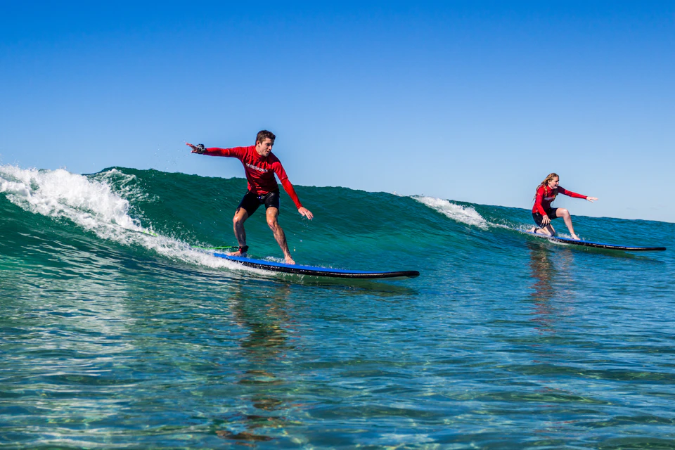 Person in a red shirt surfing on a blue surfboard, riding a wave under a clear blue sky.