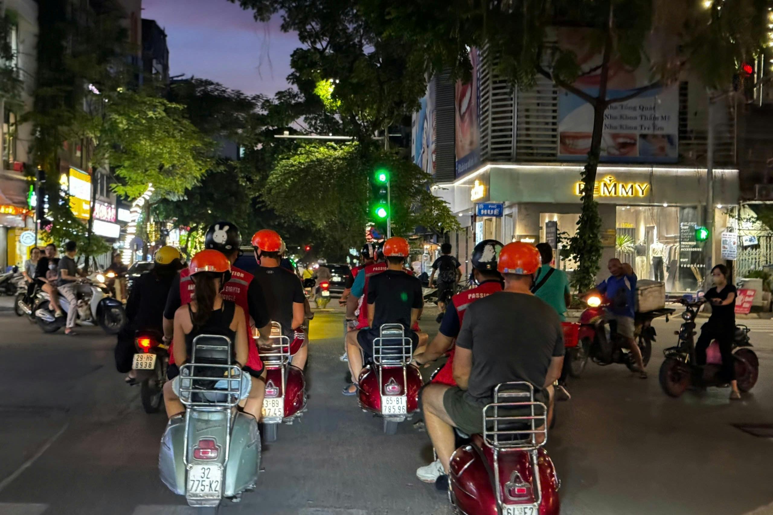 A group of people wearing helmets ride scooters at night in a city, waiting at a green traffic light.