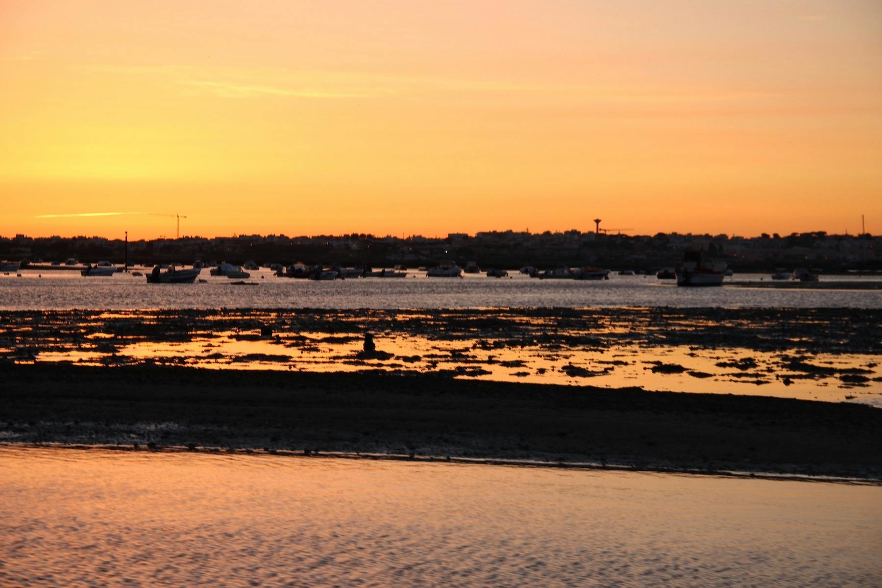 Ένα πορτοκαλί ηλιοβασίλεμα πάνω από μια παράκτια περιοχή με βάρκες στο νερό και αντανακλάσεις στην υγρή άμμο και τα ρηχά νερά