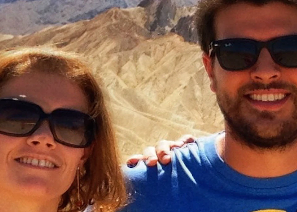 Two people wearing sunglasses standing in front of a desert landscape with rocky hills and a partly cloudy sky.
