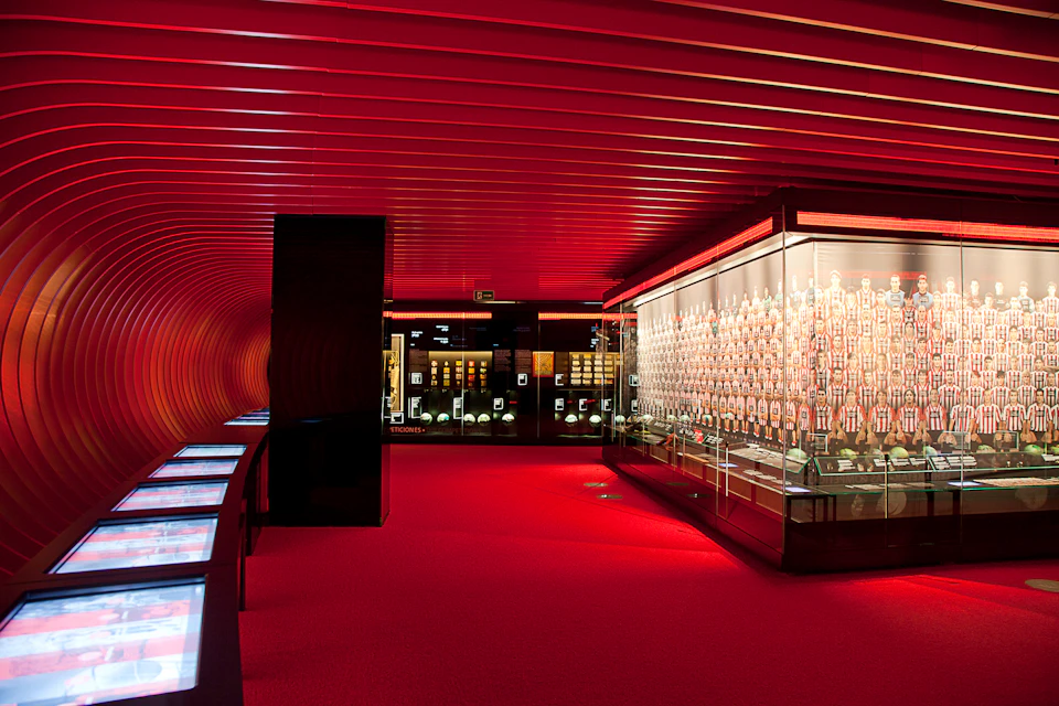 Room with red walls and carpet, showcasing sports memorabilia in illuminated glass displays, with digital information screens.