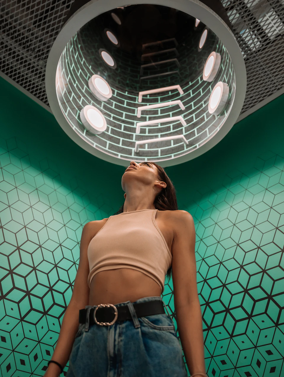 A person looks up at a cylindrical ceiling structure with lights and a brick pattern, standing in a room with green geometric walls.
