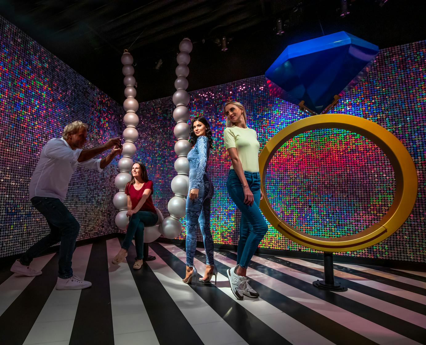 Čtyři lidé pózují pro fotografie v barevné neonově osvětlené místnosti s pruhovanou podlahou, balónkovitými sochami a velkými geometrickými tvary.