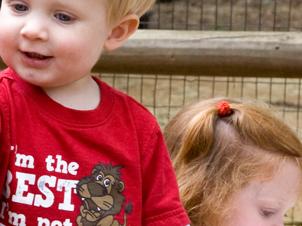 A blonde toddler in a red shirt points at a goat. A red-haired child pets another goat in a petting zoo area.