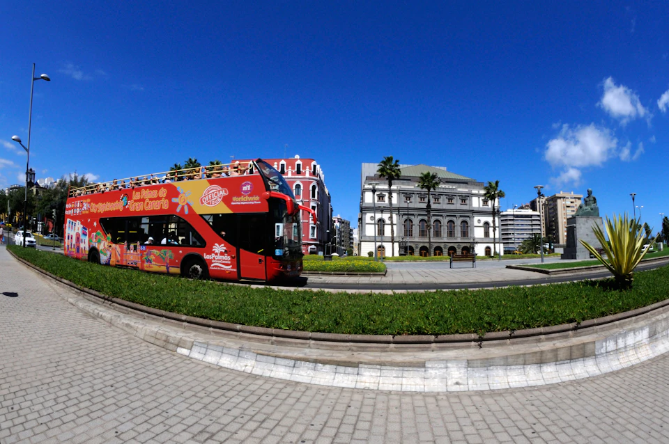A red double-decker tour bus passes by a park with green grass, palm trees, and historical buildings under a bright blue sky.