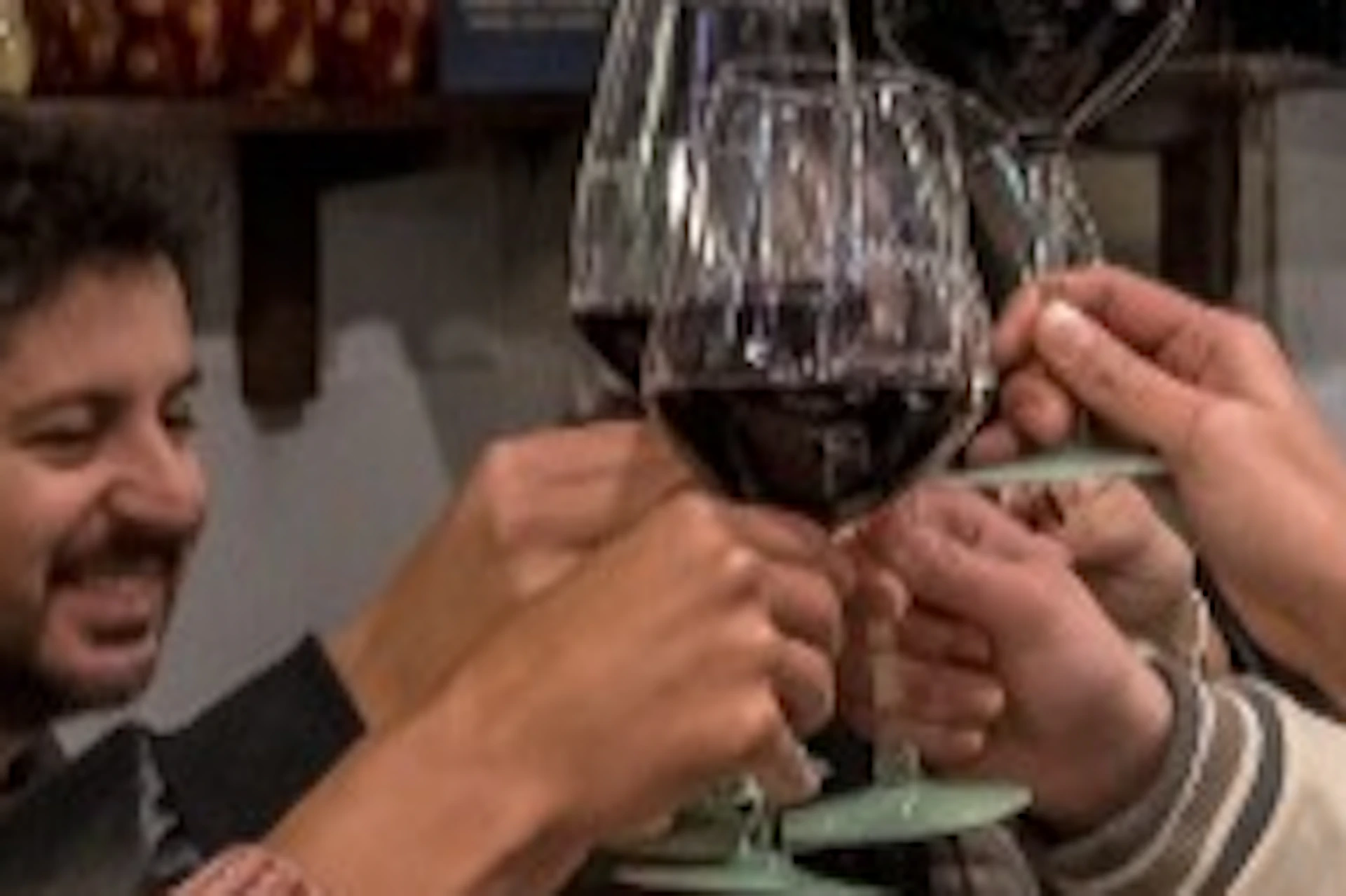 A group of people seated at a table in a cozy restaurant, toasting with glasses of red wine, surrounded by shelves of food products.