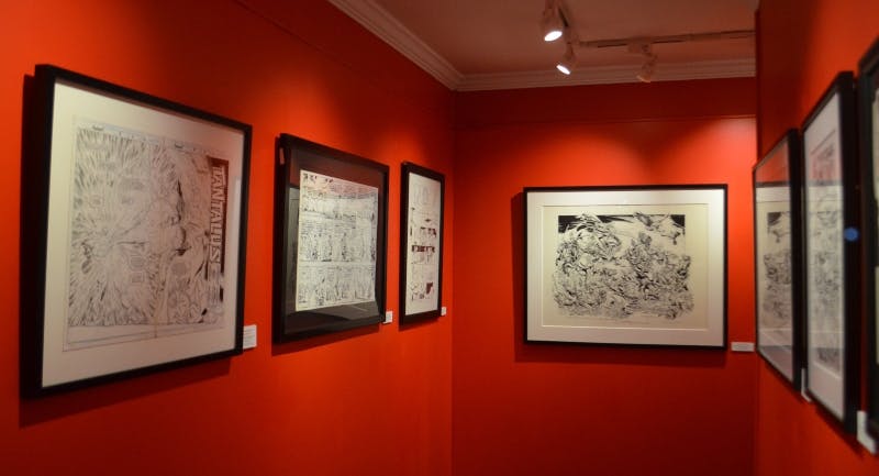 Framed black-and-white comic art displayed on bright red walls in a gallery with ceiling spotlights.