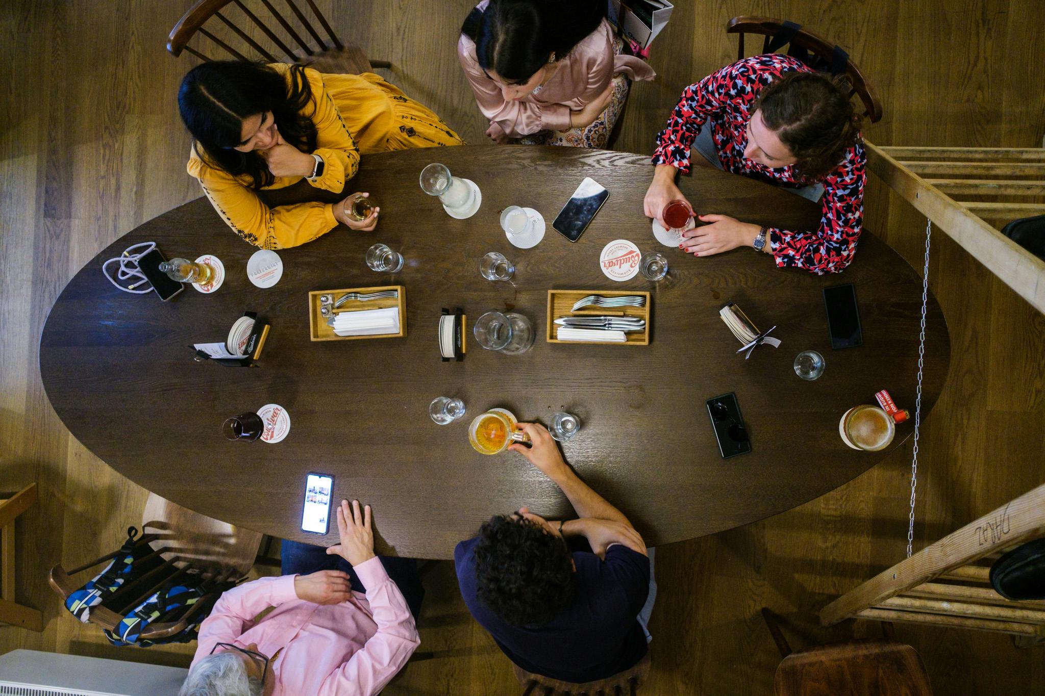 Kolem dřevěného stolu s nápoji, telefony a podnosy s jídlem sedělo pět lidí. Pohled shora.