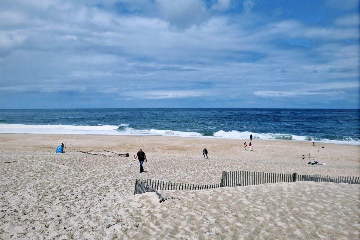 Άνθρωποι περπατούν σε μια αμμώδη παραλία δίπλα στον ωκεανό κάτω από έναν συννεφιασμένο ουρανό, με έναν μικρό περιφραγμένο αμμόλοφο σε πρώτο πλάνο.