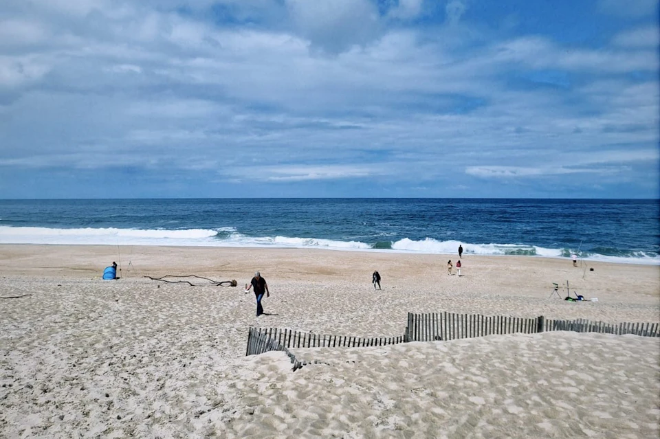 People walking on a sandy beach by the ocean under a cloudy sky, with a small fenced dune in the foreground.