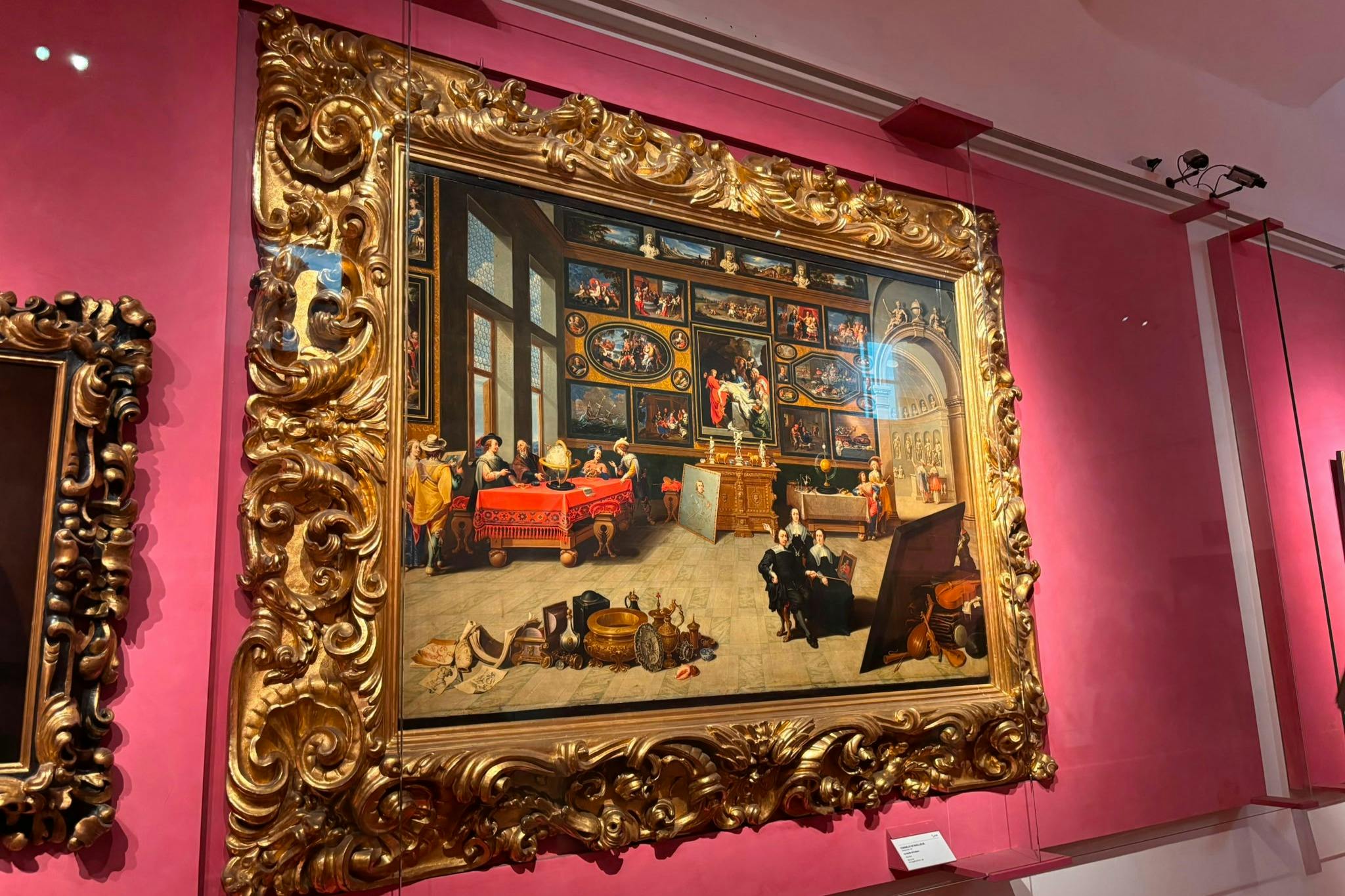 Um quadro grande, com uma moldura elaborada, retrata uma cena interior detalhada com pessoas, obras de arte e objetos, exposto numa parede cor-de-rosa.
