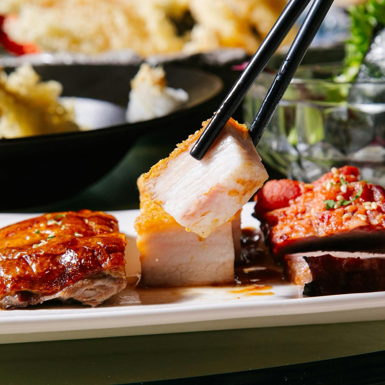 A close-up of assorted cooked meats on a plate, with chopsticks picking up a piece of crispy pork belly.