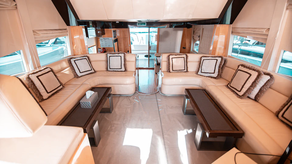 Luxurious yacht interior with beige leather couches, dark wood tables, and decorative cushions. Bright window view in the background.