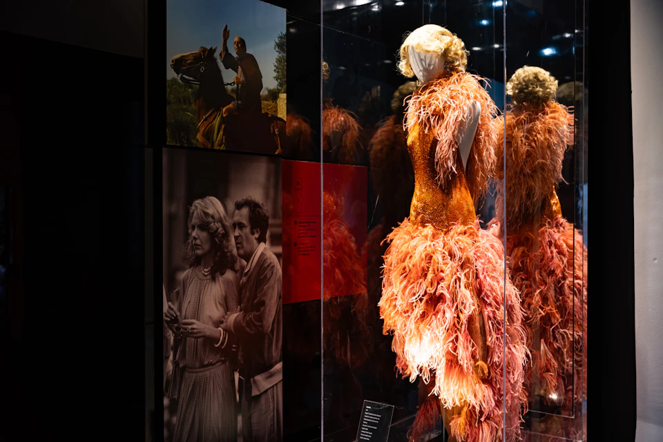 A mannequin wearing an orange feathered dress in an exhibit, with photos displayed on the wall behind it.