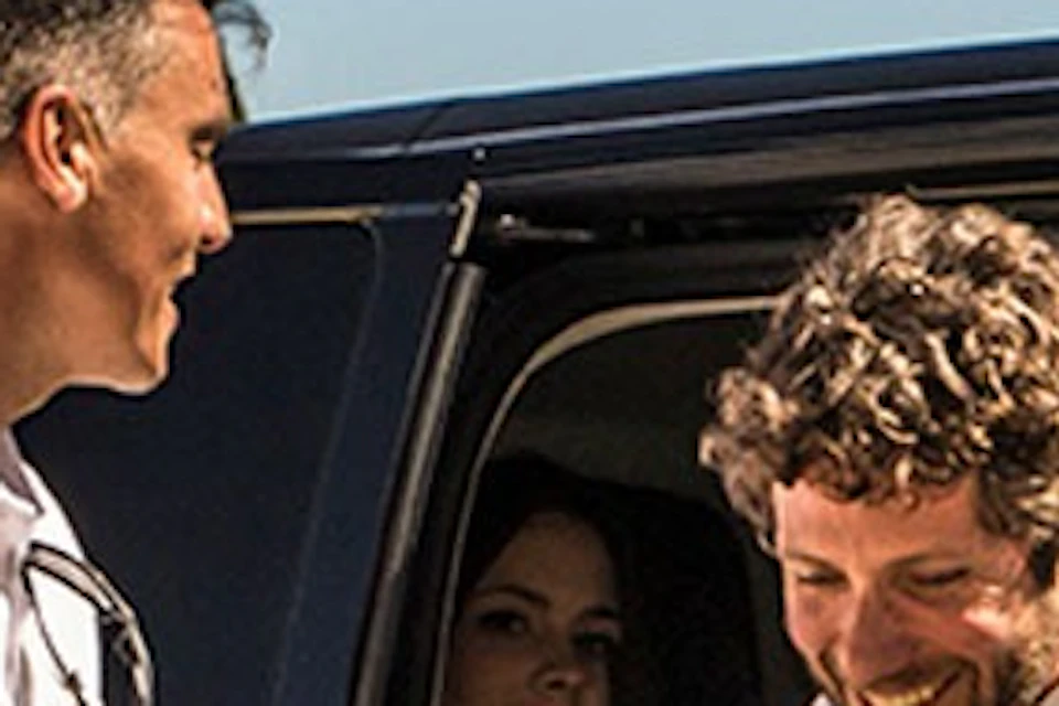 A man steps out of a black van, shaking hands with another man outside, while two other people are nearby in a sunny outdoor setting.