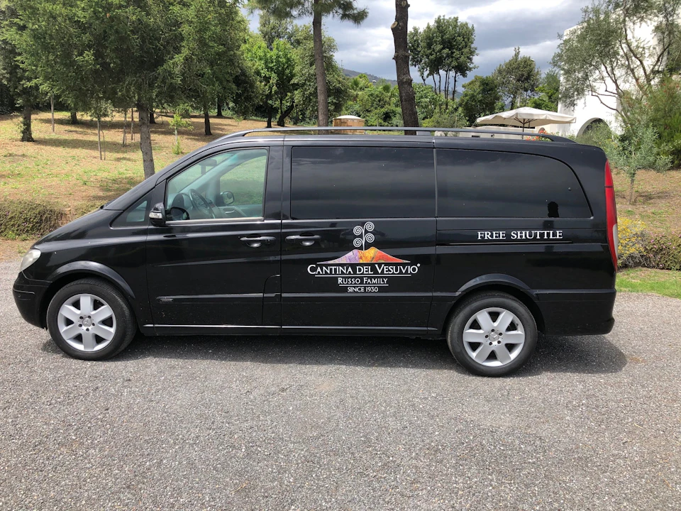 A black van labeled "Cantina del Vesuvio" with "FREE SHUTTLE" parked on a gravel road, with trees and greenery in the background.