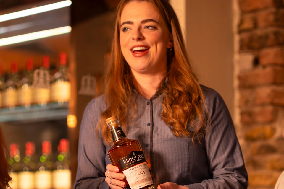 A woman standing and holding a bottle of liquor, with two people seated and glasses of liquor on a table in front of them.