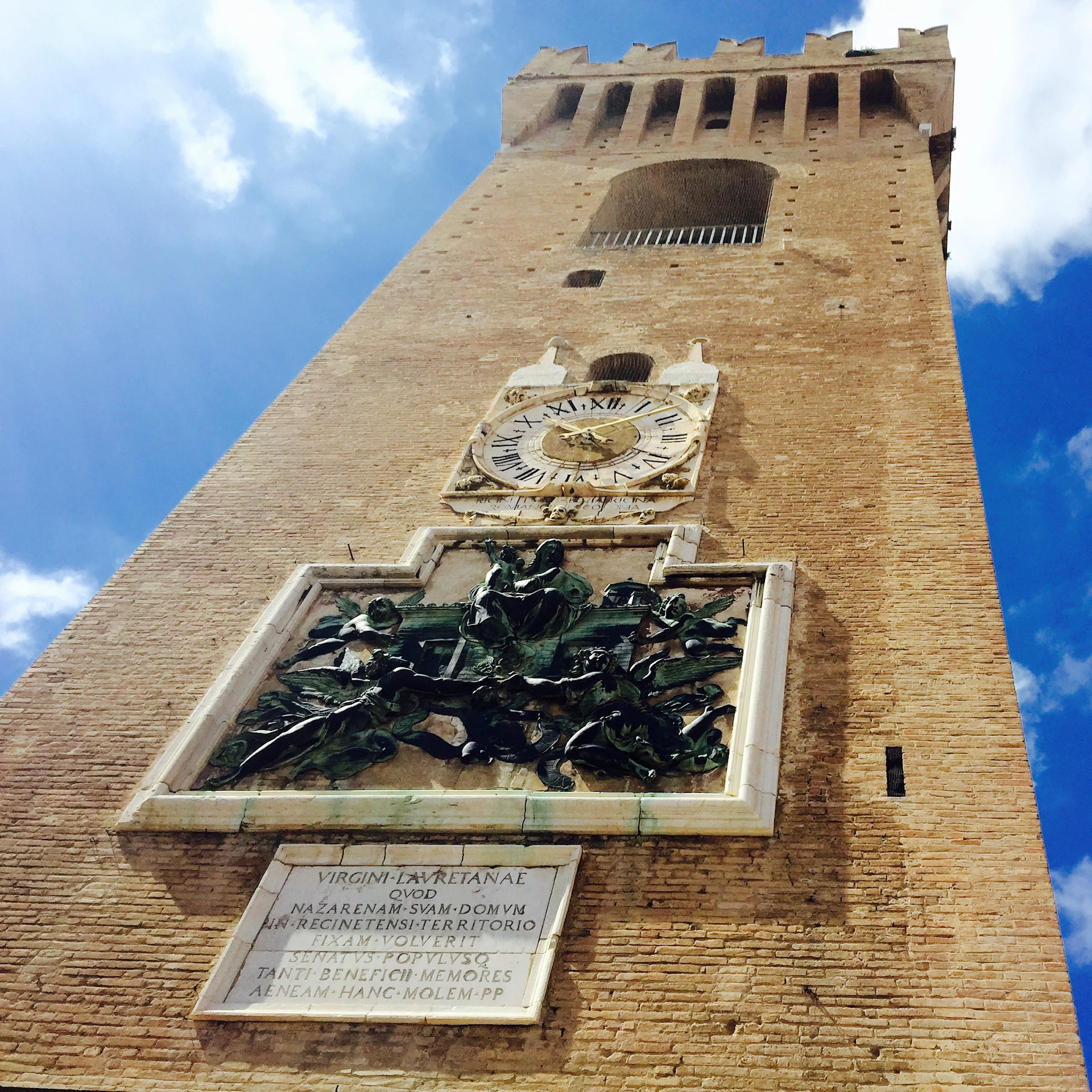 Tall brick tower with a clock, intricate sculpture, and inscription, set against a blue sky with scattered clouds.