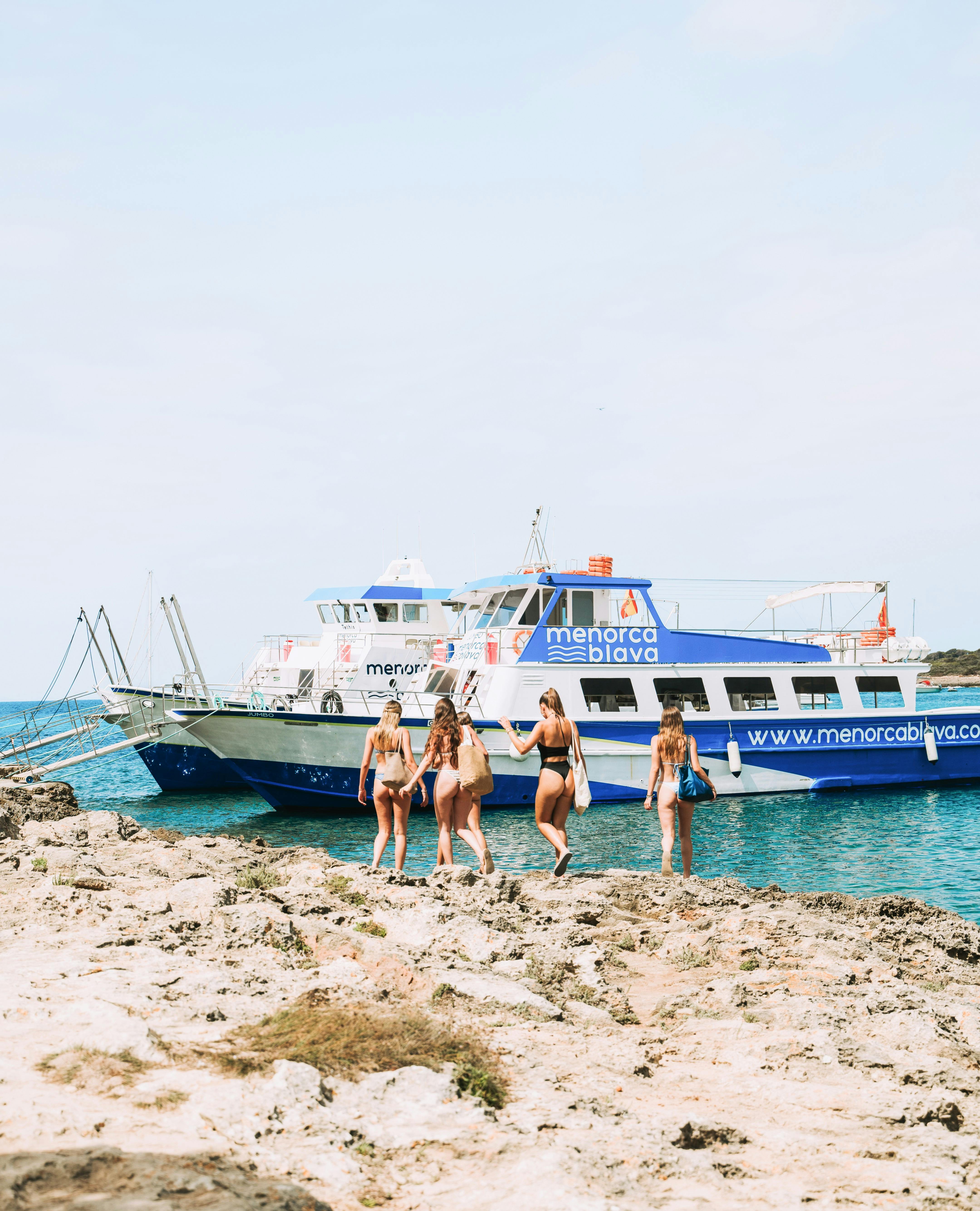 Four people in swimsuits walk towards a large boat docked by the sea with the name "Menorca Blava" visible on its side.