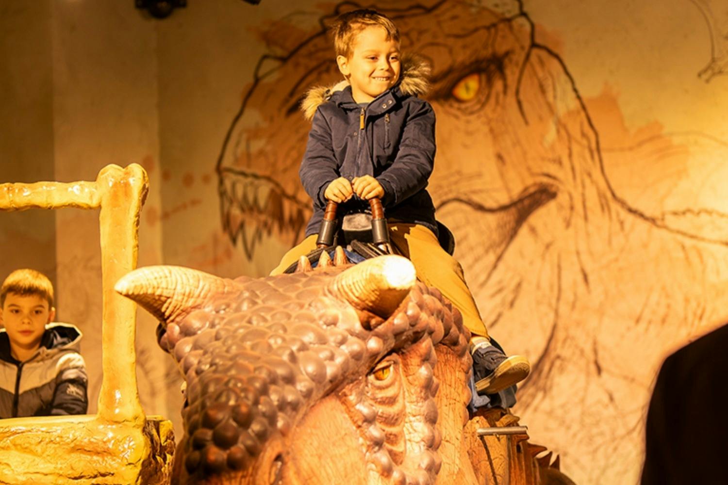 Two children in a themed play area, one riding a dinosaur figure, both enjoying the experience under warm lighting.