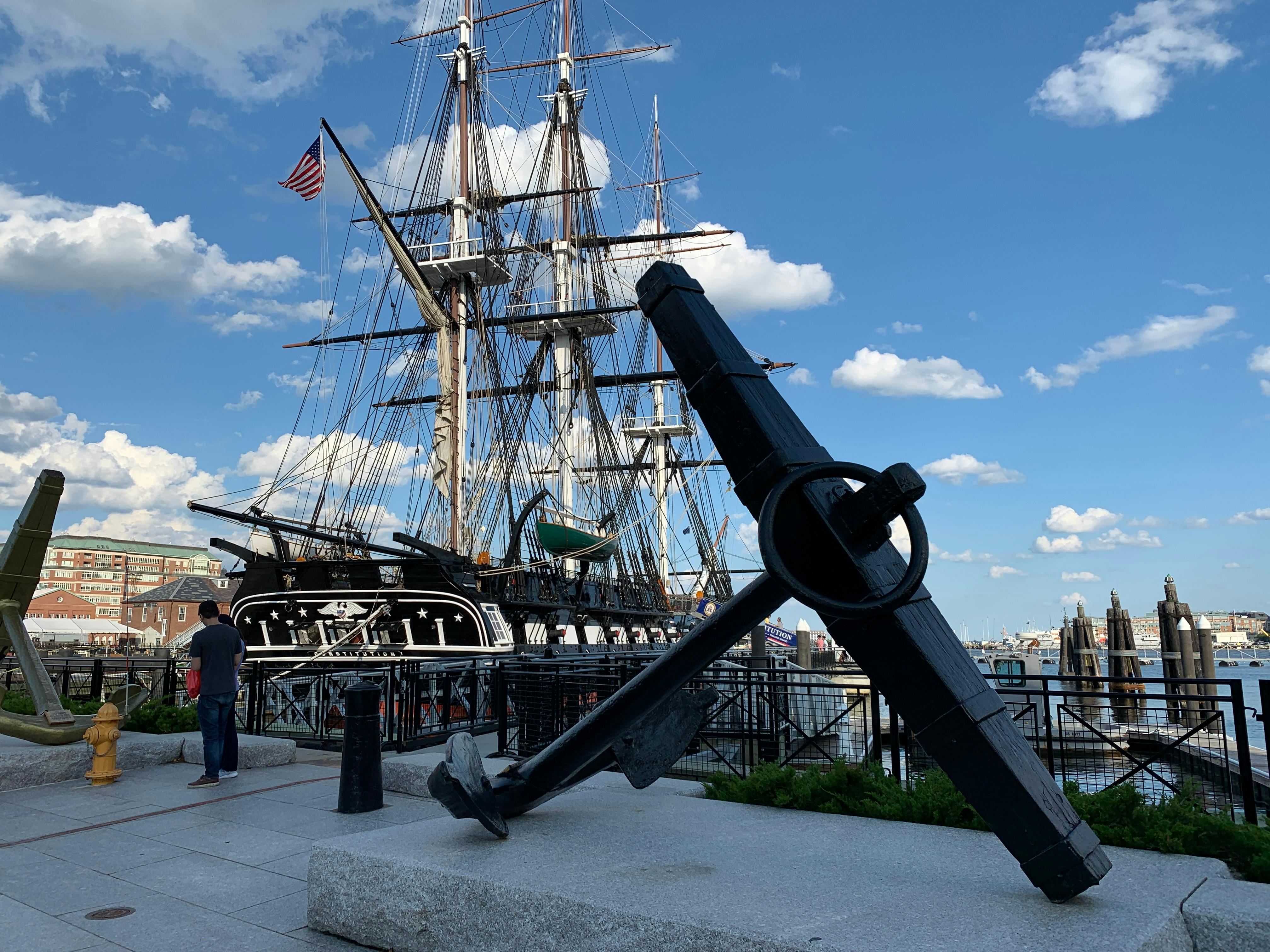 A large anchor displayed in front of a historic sailing ship with masts and rigging. A person stands nearby under a blue sky with clouds.