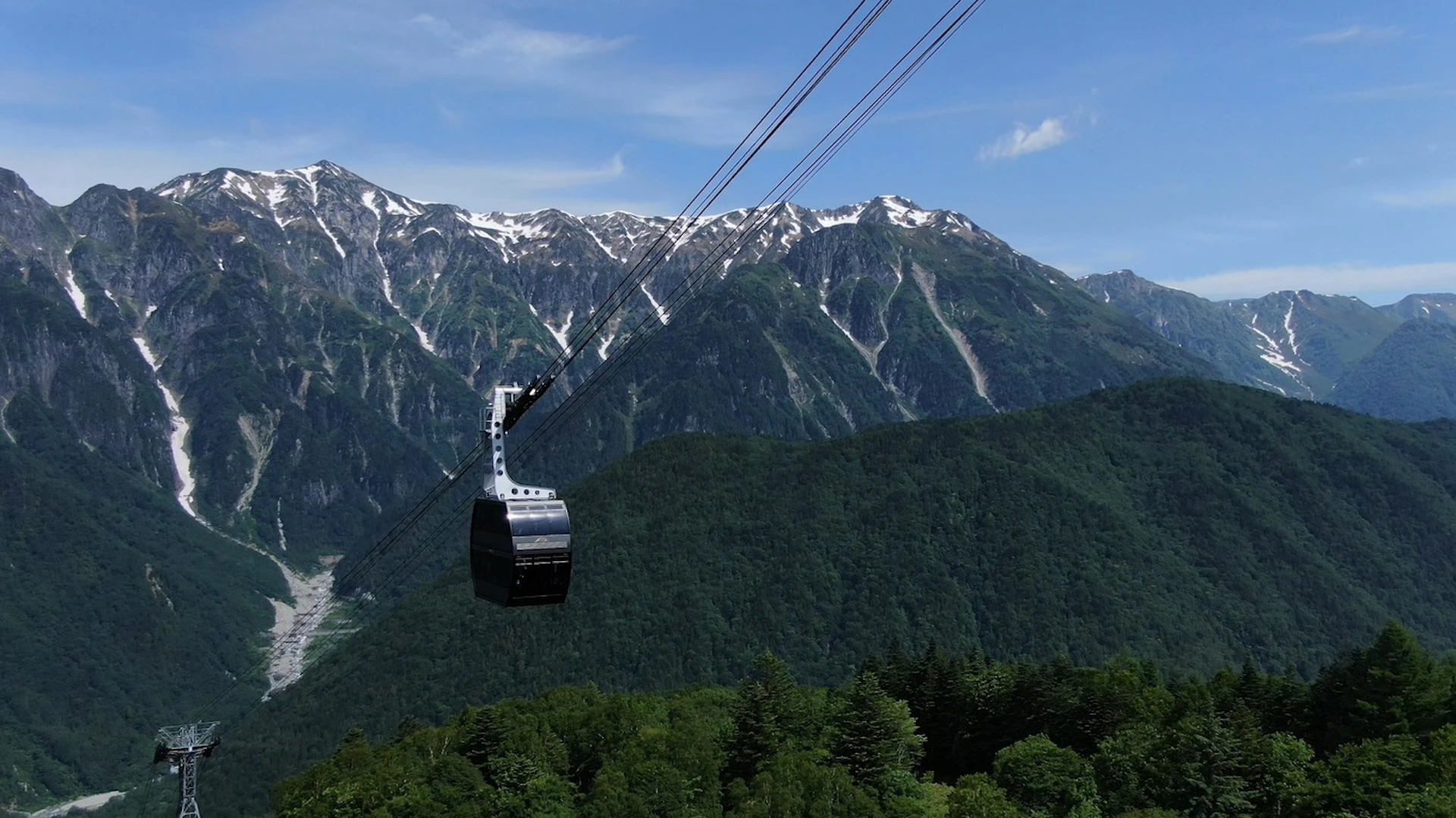 A cable car suspended on wires over a forested area with snow-capped mountains in the background under a clear sky.