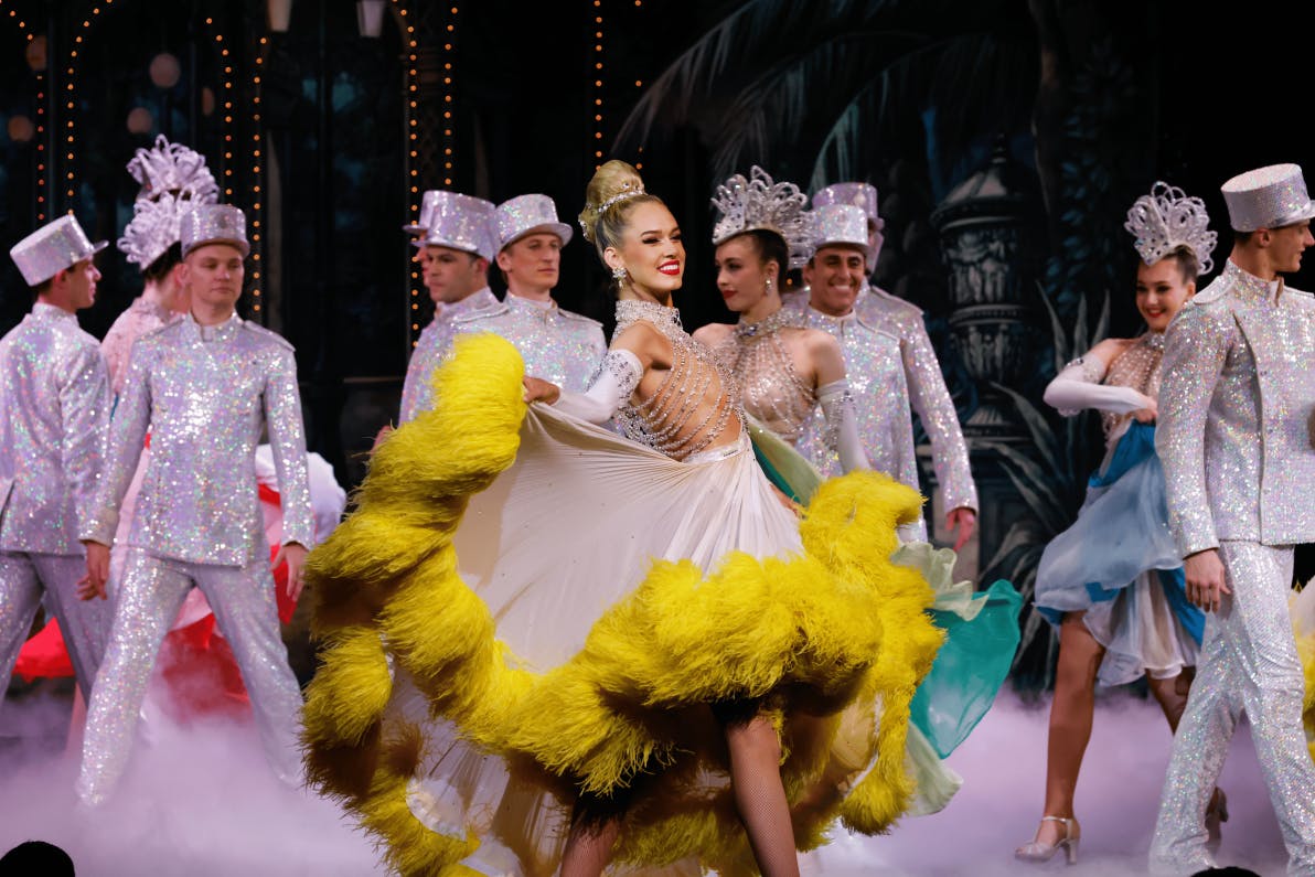 Performers in colorful costumes, including a dancer in a yellow-feathered dress, on stage with a festive backdrop.