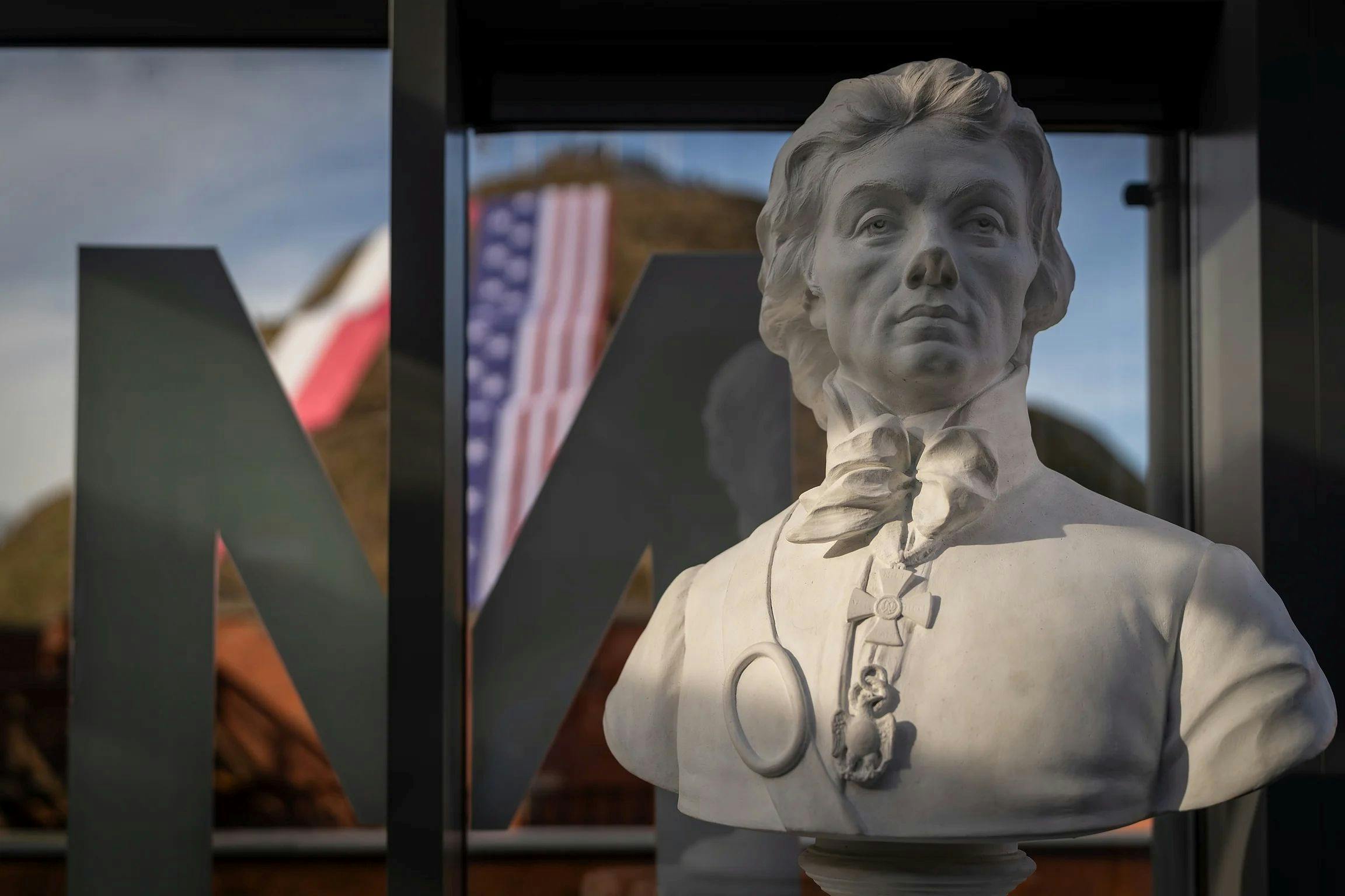 A white marble bust of a person with a bow tie and medallion, displayed indoors with a U.S. flag visible outside a window.