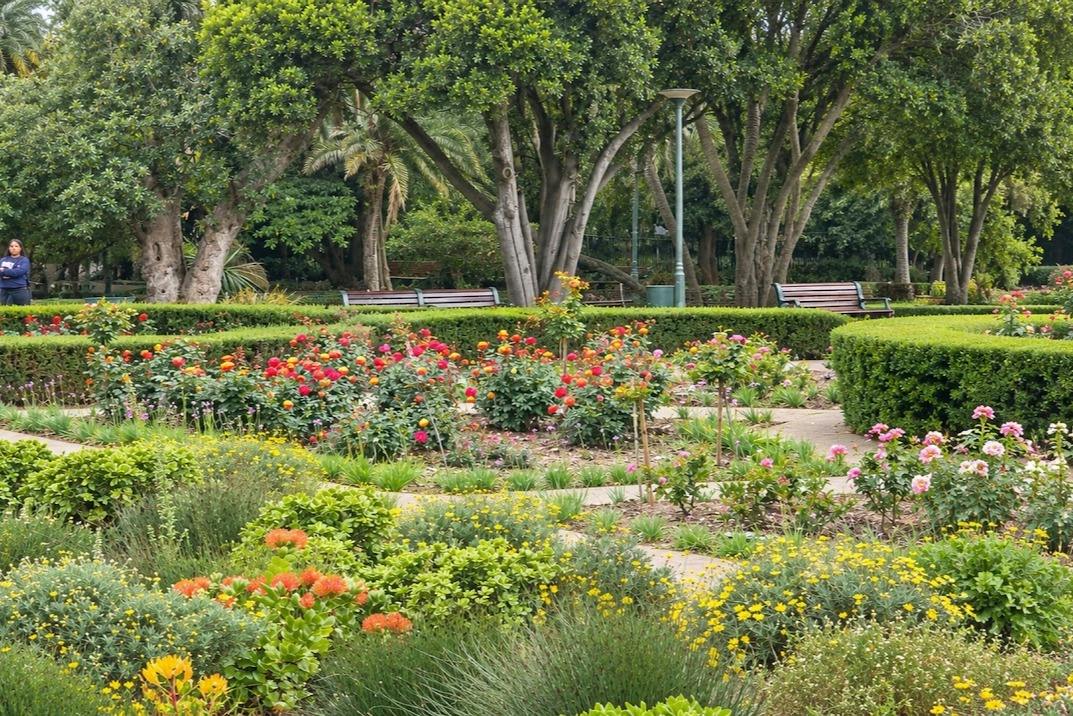 A landscaped garden with vibrant flowers, hedges, trees, and benches under a clear sky.