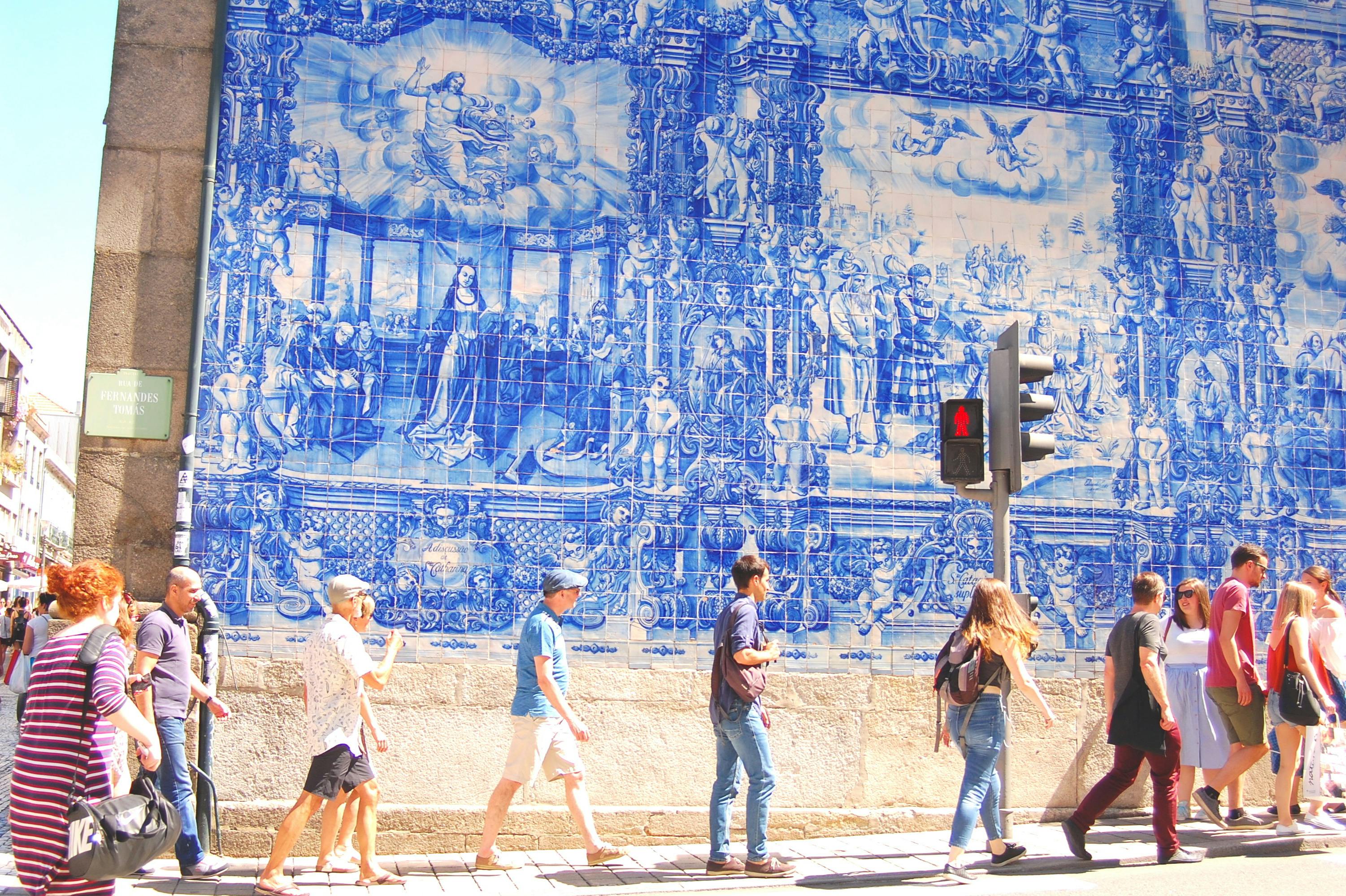 Τέσσερις άνθρωποι περνούν μπροστά από έναν μεγάλο μπλε και άσπρο τοίχο τοιχογραφίας, με ένα φανάρι που δείχνει ένα κόκκινο σήμα πεζών.