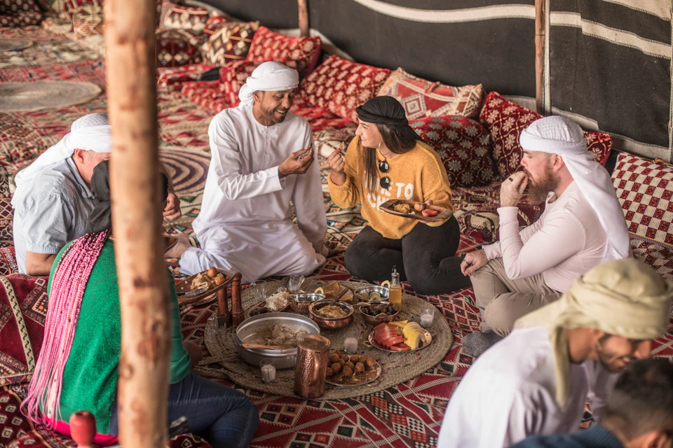 People sitting on patterned cushions, sharing and eating a meal with various dishes on a low, round table in a richly decorated tent.